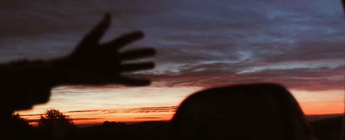 el reflejo de una mano saludando en el espejo de una furgoneta, que refleja a su vez un atardecer